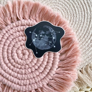 Könnte beinhalten: Ein schwarz-weißer Aufkleber mit einem Mond und Sternen in Form einer Konstellation. Der Aufkleber befindet sich auf einem rosafarbenen und weißen gehäkelten Teppich.