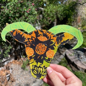 May include: A colorful, fabric goat skull with green horns and a patterned design in black, orange, and yellow. The skull is held up against a blurred green background. The number "#1" is in the top left corner.