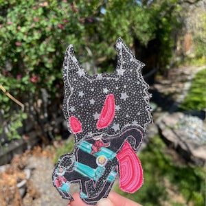 May include: A fabric patch featuring a stylized black wolf-like creature with a pink tongue and accents. The wolf's head is black with white stars. The patch has a whimsical design with a cartoonish aesthetic.
