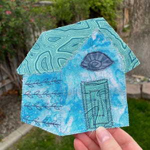 May include: A handmade fabric house with a blue and turquoise color scheme. The roof has a swirling pattern, and the body features a door, an eye-shaped design, and embroidered leaf details. The number "#1" is in the top left corner.