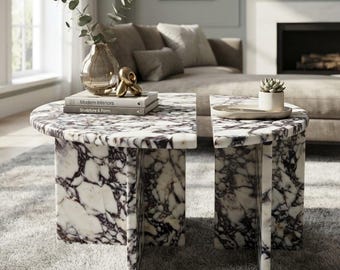 May include: Two semi-circular coffee tables made of white marble with dark purple veining. The tables feature a cross-shaped base design. On the left table, there is a glass vase with greenery and two books titled Modern Interiors and Sculpture & Form.