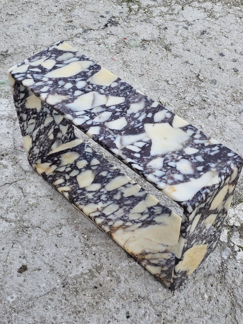 May include: A rectangular marble object with a hollow center. The marble has a pattern of white, cream, and dark purple veining. The object is sitting on a gray concrete surface.