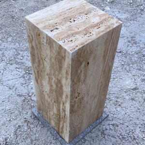 Puede incluir: Un pedestal cuadrado de piedra de color marr&oacute;n claro con una superficie rugosa y texturizada. El pedestal est&aacute; sentado sobre una superficie gris y texturizada.