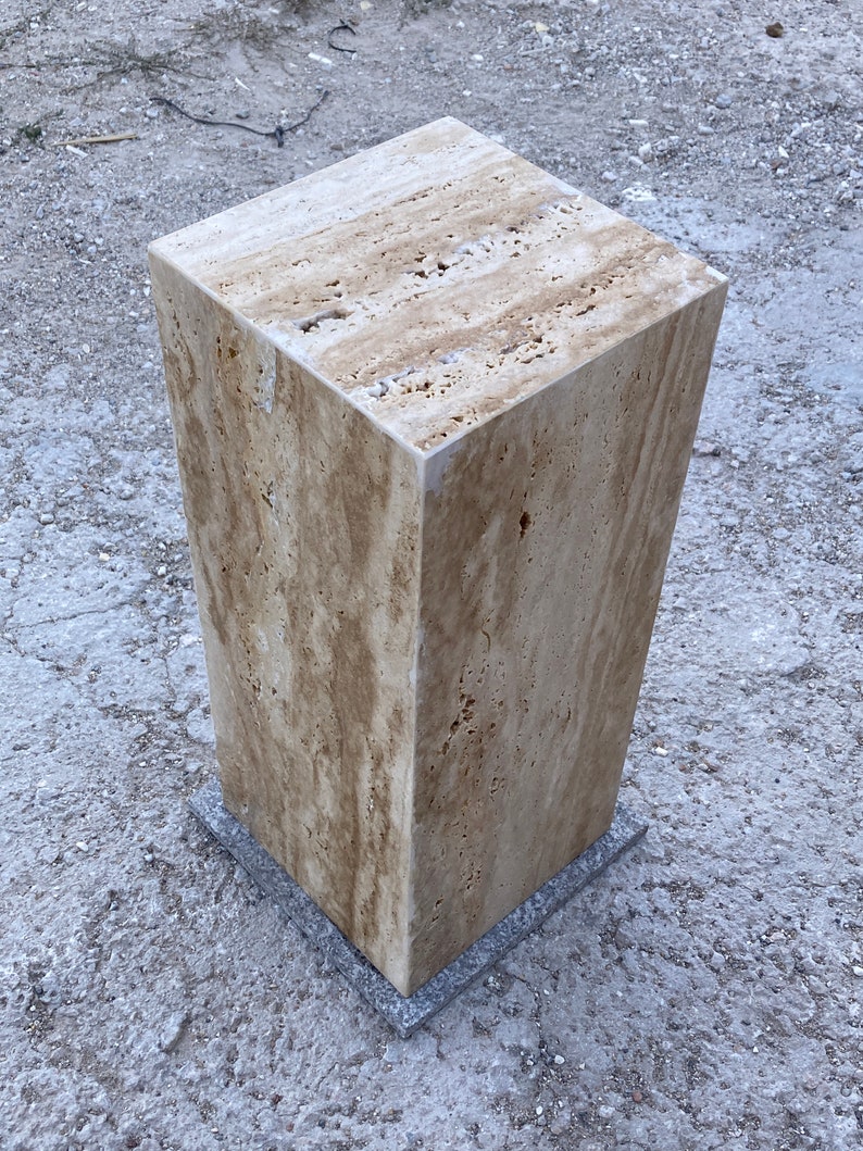 Puede incluir: Un pedestal cuadrado de piedra de color marr&oacute;n claro con una superficie rugosa y texturizada. El pedestal est&aacute; sentado sobre una superficie gris y texturizada.