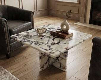 May include: A square coffee table featuring a white marble surface with prominent dark purple and black veining, supported by a matching cross-shaped base. The table holds a small silver bowl filled with nuts, a thick book, and a ceramic vase.
