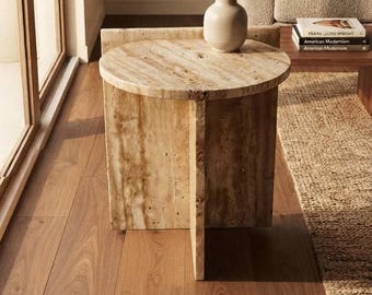 May include: A round side table constructed from beige travertine stone with a natural porous texture. The table features a circular top supported by a cross-shaped base made of two intersecting vertical panels of the same stone material.