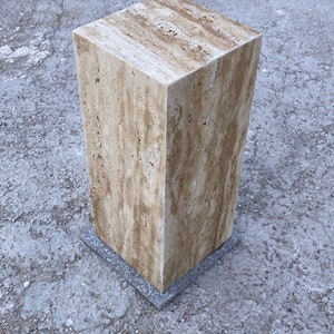 Puede incluir: Un pedestal cuadrado de piedra travertina de color marr&oacute;n claro con una base gris. El pedestal est&aacute; sobre una superficie de hormig&oacute;n gris.