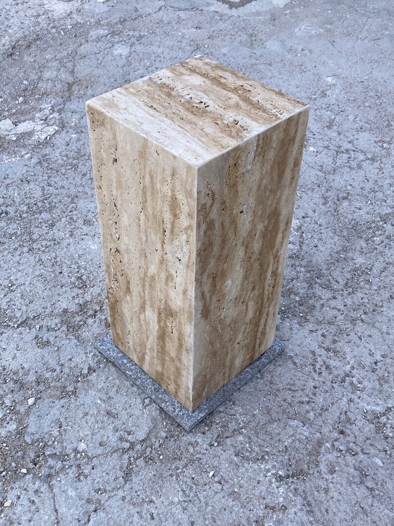 Puede incluir: Un pedestal cuadrado de piedra travertina de color marr&oacute;n claro con una base gris. El pedestal est&aacute; sobre una superficie de hormig&oacute;n gris.