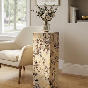 May include: A tall, rectangular pedestal with a polished, cream-colored surface featuring dark brown and black veining throughout. A clear glass vase containing a bouquet of dried eucalyptus stems and light-colored roses sits on top.