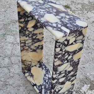 May include: A rectangular marble side table with a unique U-shaped design. The table features a cream, black, and tan patterned surface. The table is made of stone and is suitable for indoor or outdoor use.