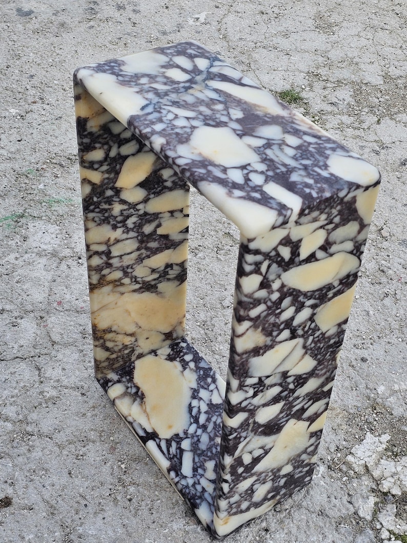 May include: A rectangular marble side table with a unique U-shaped design. The table features a cream, black, and tan patterned surface. The table is made of stone and is suitable for indoor or outdoor use.