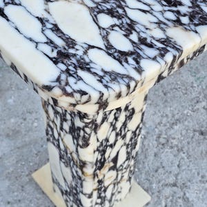 K&ouml;nnte beinhalten: Ein Marmor-Sockeltisch mit quadratischer Platte und rechteckigem Sockel. Der Tisch weist ein auff&auml;lliges Muster aus wei&szlig;en und schwarzen Adern im gesamten Stein auf. Der Sockel hat eine S&auml;ulenform.