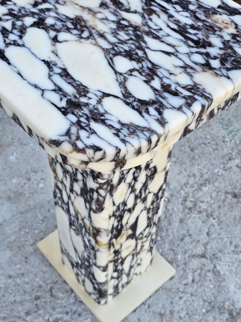K&ouml;nnte beinhalten: Ein Marmor-Sockeltisch mit quadratischer Platte und rechteckigem Sockel. Der Tisch weist ein auff&auml;lliges Muster aus wei&szlig;en und schwarzen Adern im gesamten Stein auf. Der Sockel hat eine S&auml;ulenform.