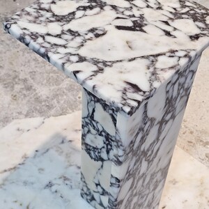 May include: A square marble pedestal table with a white and dark gray veined pattern. The table has a rectangular base and a square top. The marble has a polished finish.