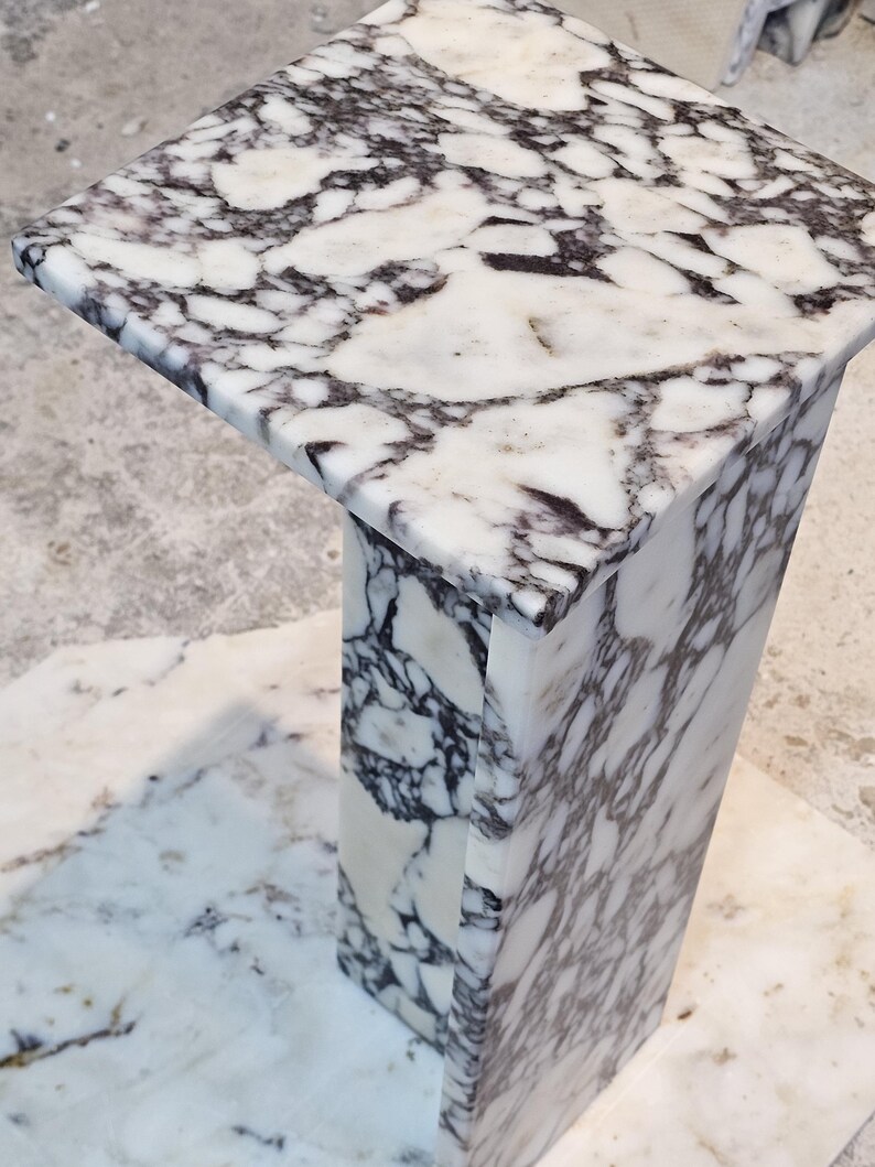 May include: A square marble pedestal table with a white and dark gray veined pattern. The table has a rectangular base and a square top. The marble has a polished finish.