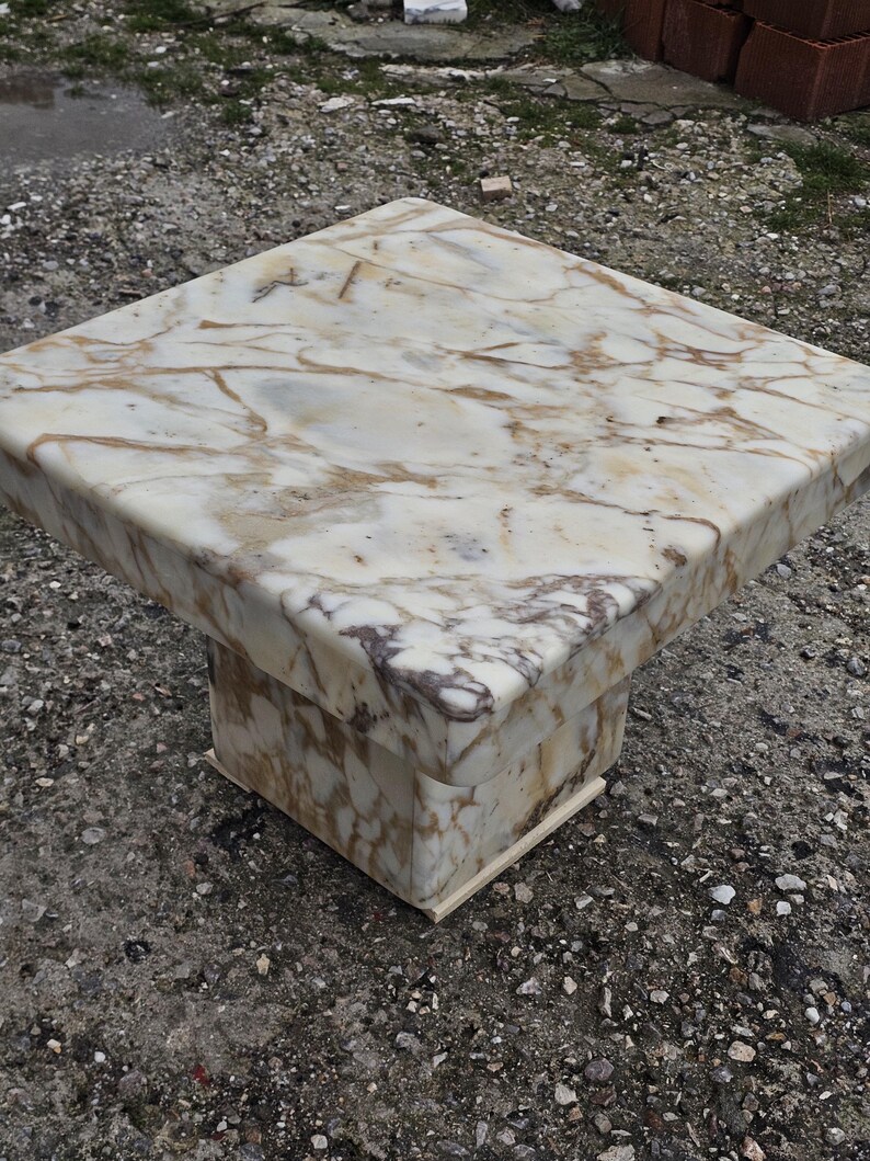 May include: A square side table constructed from white marble with prominent brown and grey veining. The table features a thick, flat top surface supported by a central, block-style pedestal base that matches the marble pattern of the top.