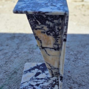 May include: A small, marble side table with a unique, angular design. The table features a rectangular top and base, connected by a triangular support. The marble has a cream background with dark blue and black veining.