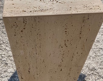 May include: A beige, rectangular stone pedestal with a textured surface. The pedestal has a trapezoidal shape, wider at the top and tapering towards the base. The stone has small holes and imperfections.