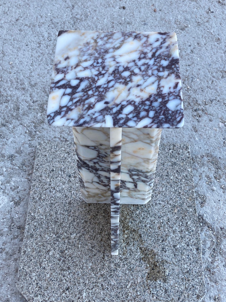 May include: A square pedestal stand crafted from polished marble featuring a distinct veined pattern in shades of white, cream, and deep purple. The structure consists of a flat top surface supported by a central vertical column base.