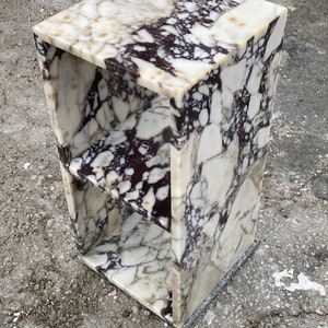 May include: A white and brown marble end table with two shelves. The table has a square top and a rectangular base.