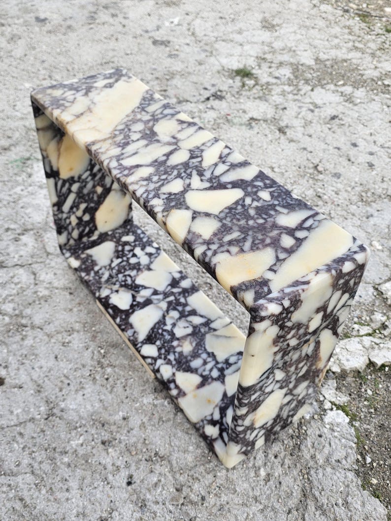 May include: A rectangular marble shelf with a unique open design. The shelf features a cream, black, and gray speckled pattern. The shelf is made of stone and is suitable for displaying decorative items.