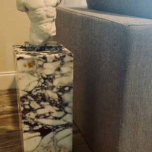 May include: A white marble end table with a white plaster sculpture of a torso on top. The table has a brown and white marbled pattern. The table is next to a gray couch.