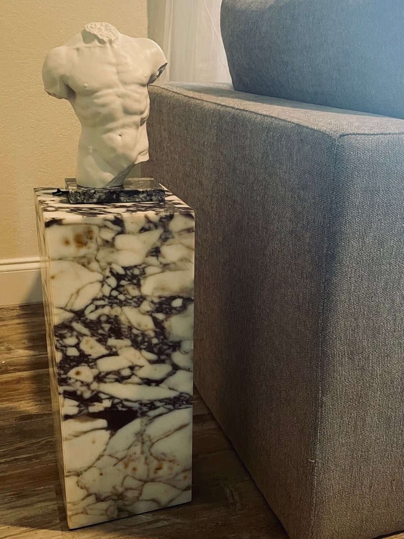 May include: A white marble end table with a white plaster sculpture of a torso on top. The table has a brown and white marbled pattern. The table is next to a gray couch.