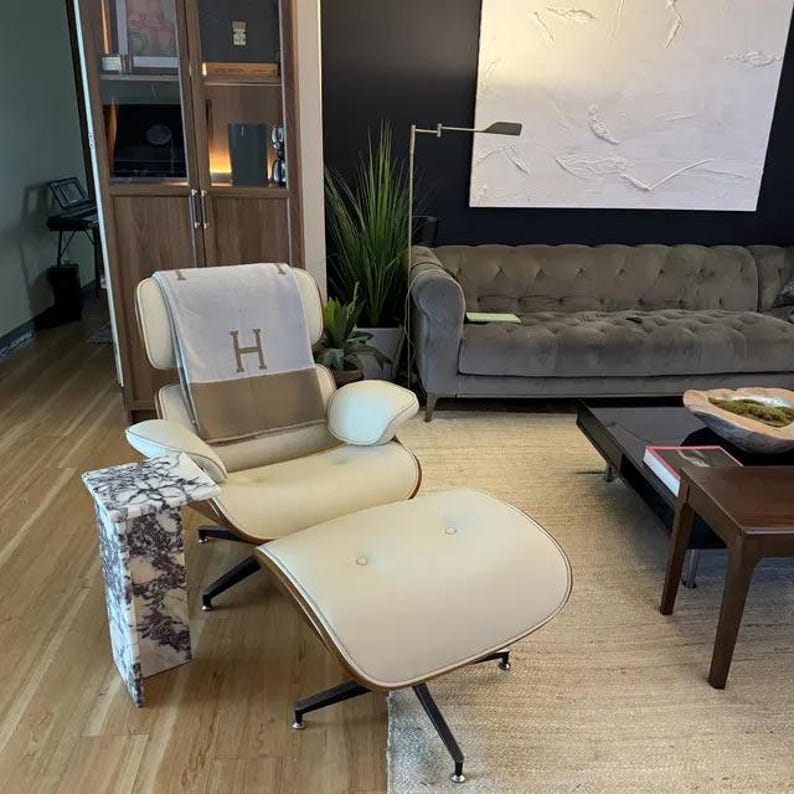May include: A cream-colored lounge chair with a matching ottoman and a marble side table. The chair has a brown trim and a blanket with the letter "H" draped over it. The chair and ottoman sit on a light brown rug.