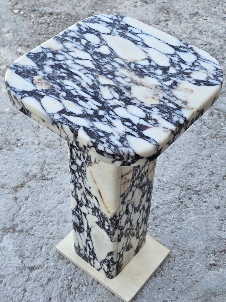 K&ouml;nnte beinhalten: Ein Marmorsockel mit quadratischer Oberseite und rechteckiger Basis. Die Oberseite weist ein wei&szlig;es und schwarzes Marmormuster auf. Die S&auml;ule des Sockels ist ebenfalls aus Marmor mit einem &auml;hnlichen Muster. Die Basis ist hellbeige.