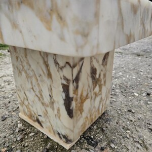 May include: A square pedestal table crafted from polished white marble featuring prominent golden-brown and dark grey veining throughout the stone surface. The structure consists of a thick, flat top supported by a solid, block-shaped base.