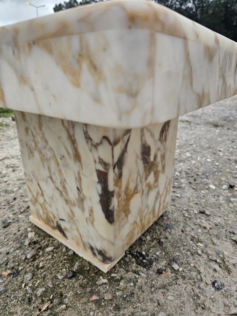 May include: A square pedestal table crafted from polished white marble featuring prominent golden-brown and dark grey veining throughout the stone surface. The structure consists of a thick, flat top supported by a solid, block-shaped base.