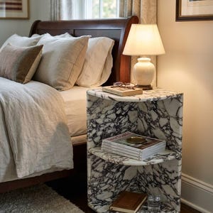 May include: A corner bedside table crafted from white marble with prominent dark grey veining. The unit features two open shelves, with the top surface holding a lamp, a book, and glasses, while the middle shelf holds magazines and the bottom holds a book.