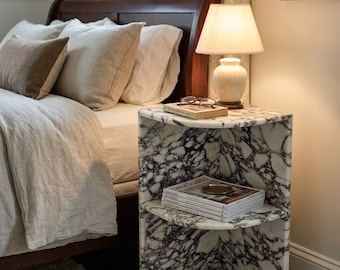 May include: A corner nightstand crafted from white marble with prominent dark grey veining. The unit features two open shelves, with the top surface holding a lamp, a book, and glasses, while the middle shelf holds magazines and the bottom holds a book.