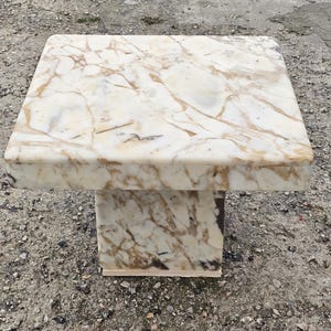 May include: A square pedestal stand crafted from polished marble featuring a cream-colored base with prominent tan and brown veining throughout the surface. The structure consists of a flat top supported by a single, thick, central rectangular column.