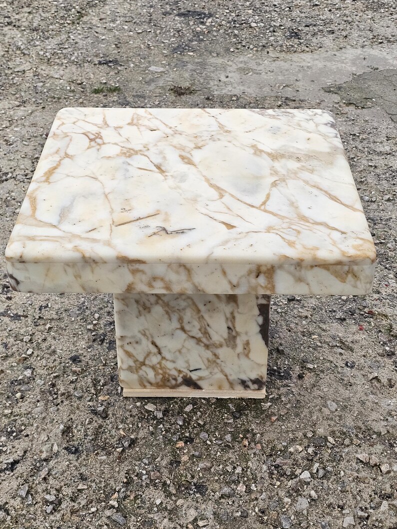 May include: A square pedestal stand crafted from polished marble featuring a cream-colored base with prominent tan and brown veining throughout the surface. The structure consists of a flat top supported by a single, thick, central rectangular column.