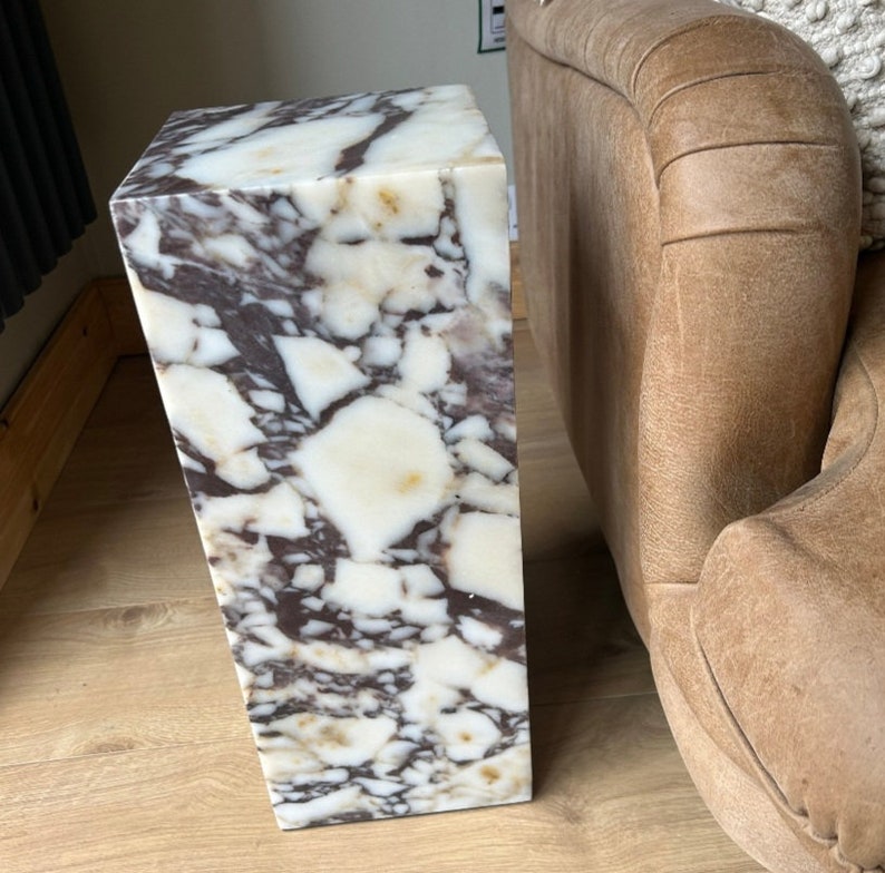 May include: A white marble pedestal with brown and gray veining. The pedestal is rectangular and has a smooth, polished surface.