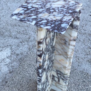 May include: A pedestal stand crafted from white and dark purple veined marble. The structure features a flat square top surface supported by a vertical column base with a distinct angular design, showcasing natural stone patterns throughout the piece.