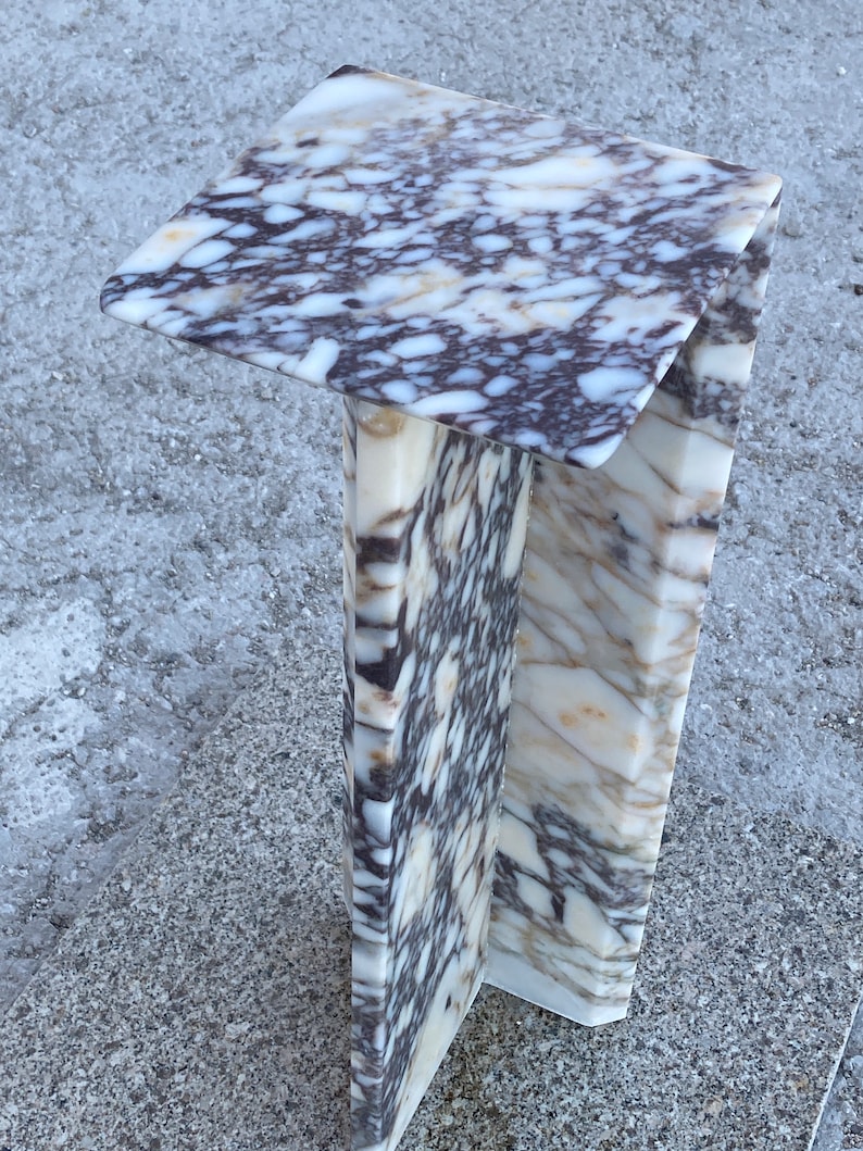 May include: A pedestal stand crafted from white and dark purple veined marble. The structure features a flat square top surface supported by a vertical column base with a distinct angular design, showcasing natural stone patterns throughout the piece.