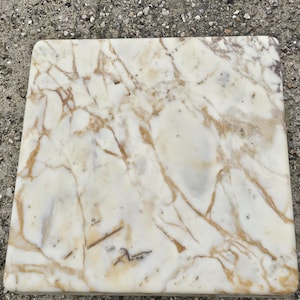 May include: A square slab of polished white marble featuring prominent, irregular veining in shades of tan, gold, and light brown. The surface is smooth with rounded edges, displaying a natural stone pattern that extends across the entire top face.