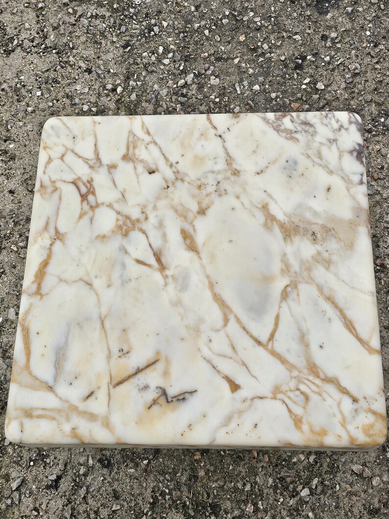 May include: A square slab of polished white marble featuring prominent, irregular veining in shades of tan, gold, and light brown. The surface is smooth with rounded edges, displaying a natural stone pattern that extends across the entire top face.