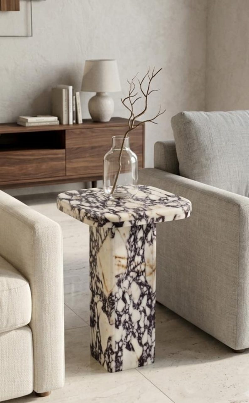 May include: A square side table crafted from white and purple veined marble with a thick pedestal base. A clear glass vase containing a single thin, bare branch sits on the flat top surface of the table, which is positioned between two upholstered chairs.