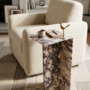 May include: A C-shaped side table crafted from white and deep purple veined marble. The table surface holds a small ceramic mug, a brown leather-bound notebook with a pen resting on top, and a small, standing brass bird figurine.