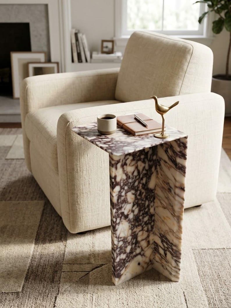 May include: A C-shaped side table crafted from white and deep purple veined marble. The table surface holds a small ceramic mug, a brown leather-bound notebook with a pen resting on top, and a small, standing brass bird figurine.