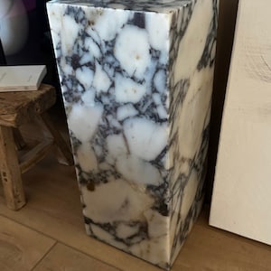May include: A white marble pedestal with gray and brown veining. The pedestal is square and has a smooth, polished surface.