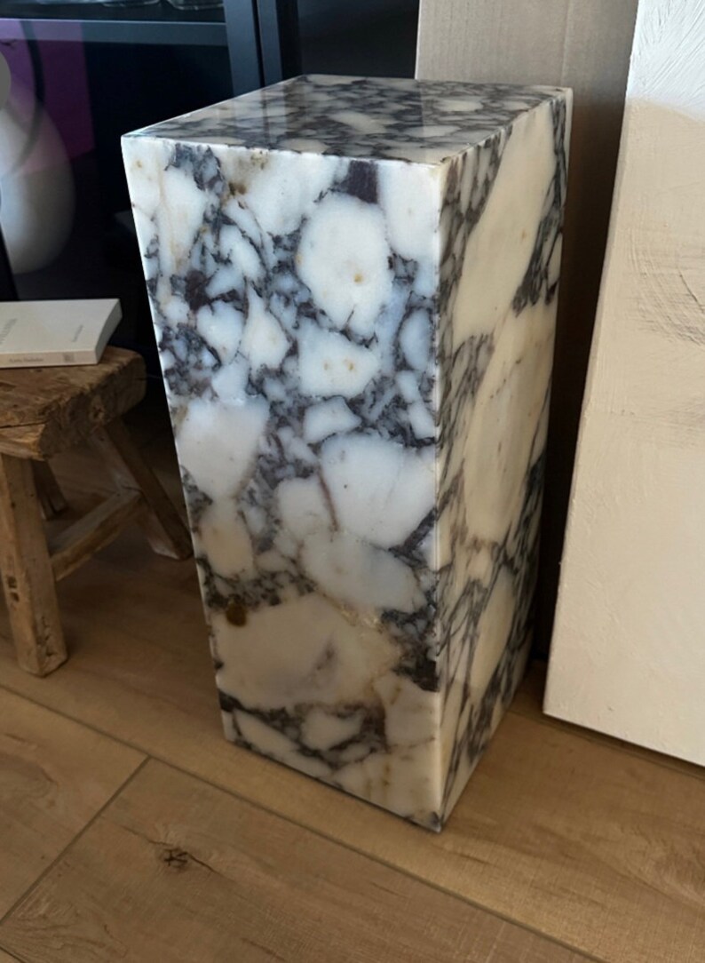 May include: A white marble pedestal with gray and brown veining. The pedestal is square and has a smooth, polished surface.