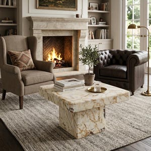 May include: A square coffee table made of polished cream and brown veined stone with a thick block base. On the table sits a small potted green plant, a clear glass tumbler on a gold tray, and two stacked books titled Kinfolk and Architectural.