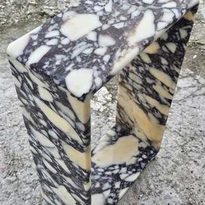 May include: A rectangular marble sculpture with a unique open-frame design. The stone features a striking pattern of black, white, and cream-colored veining. The sculpture's smooth, polished surface highlights the natural beauty of the marble.