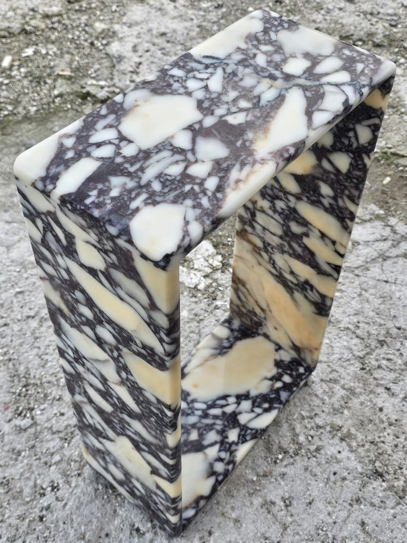 May include: A rectangular marble sculpture with a unique open-frame design. The stone features a striking pattern of black, white, and cream-colored veining. The sculpture's smooth, polished surface highlights the natural beauty of the marble.