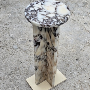 Può includere: Un tavolino a piedistallo in marmo con piano rotondo e base a colonna sfaccettata. Il piano presenta un motivo marmorizzato bianco e grigio scuro. La base è sostenuta da una base quadrata in legno chiaro. Ideale per esporre oggetti decorativi.