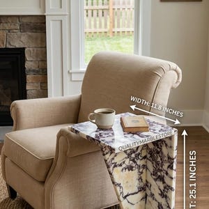 May include: A C-shaped side table made of white and purple veined marble. The table features the text QUALITYMARBLESTORE on its side. It measures 11.8 inches in width and 25.1 inches in height, holding a ceramic mug and a small book on its surface.